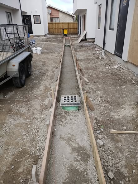 Shallow channel with aggregate base and sump grate, timber edging, at a Christchurch townhouse site — drainage and site preparation.