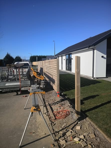 Horizontal-slat timber fence under construction with a yellow mitre saw, trailer and new lawn — fencing in Christchurch.