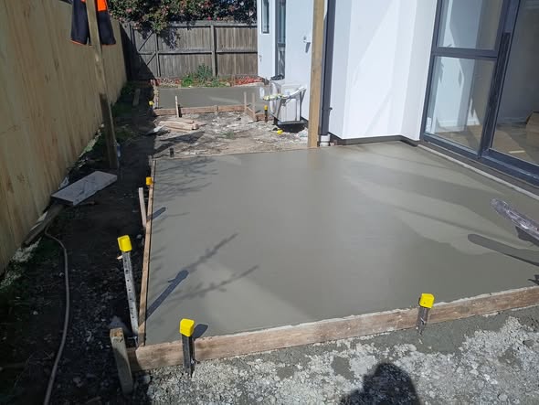 Large fresh concrete patio with yellow-capped formwork pegs beside ranch sliders and a new timber fence.